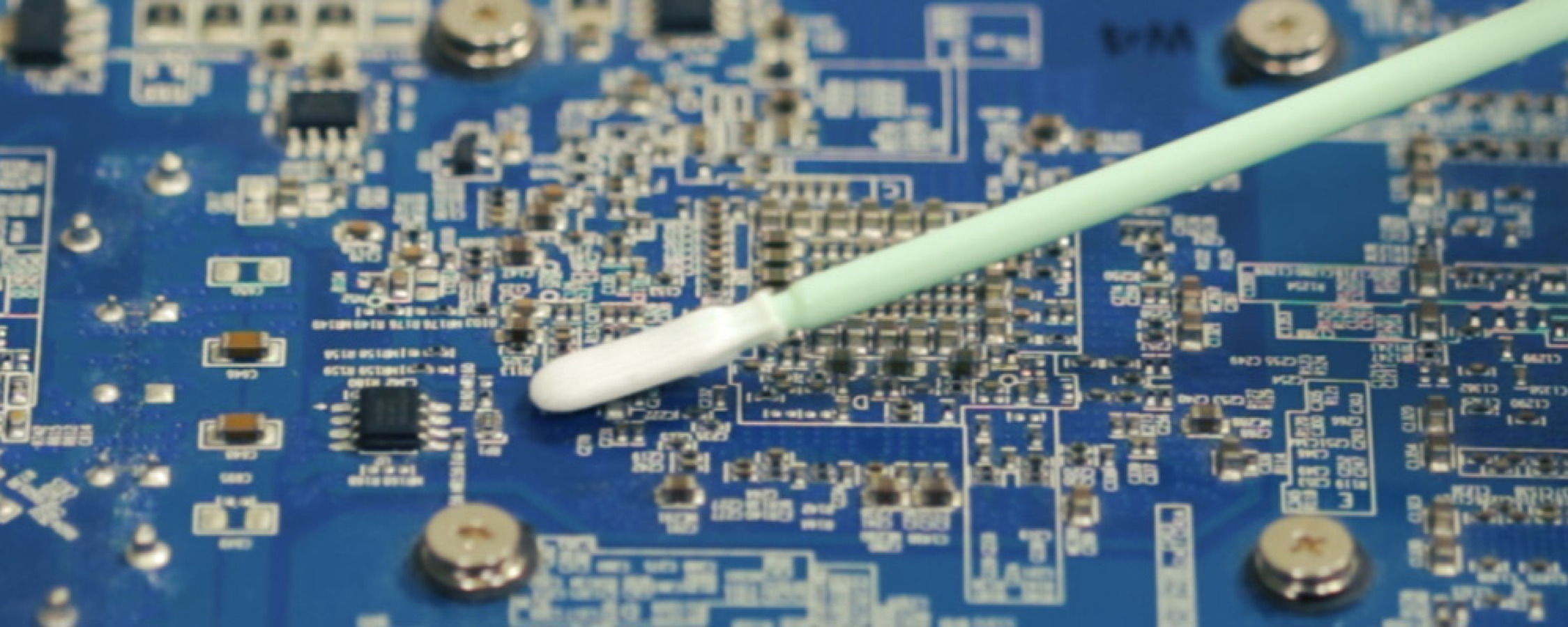 A close up of a Q+ swab removing dust from a blue circuit board